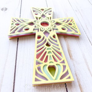 May include: A layered paper cross with a mandala design in yellow, green, pink, and purple. The cross is made of multiple layers of paper that are cut out to create a delicate and intricate design.