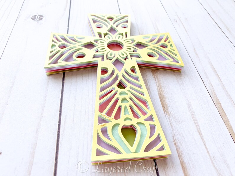 May include: A layered paper cross with a mandala design in yellow, green, pink, and purple. The cross is made of multiple layers of paper that are cut out to create a delicate and intricate design.