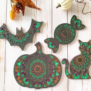 Puede incluir: Cuatro decoraciones de Halloween de madera en capas con diseños de mandalas. Las decoraciones son un murciélago, una calabaza, un caramelo y un gato. Las decoraciones están pintadas en tonos de verde, naranja y morado.