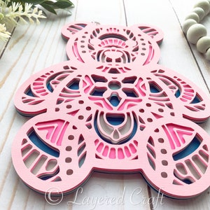 May include: A layered papercraft bear ornament with a pink, blue, and gray mandala design. The bear is cut out of paper and layered to create a 3D effect.