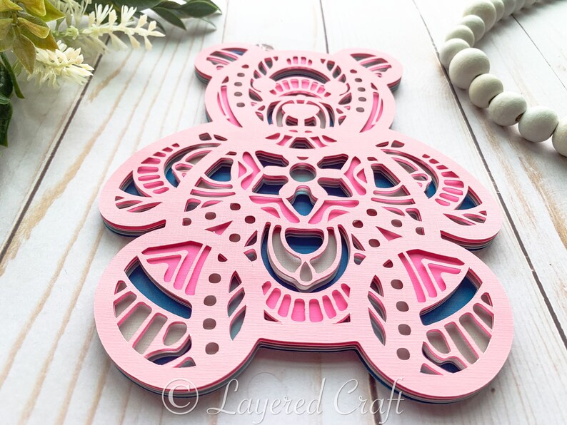 May include: A layered papercraft bear ornament with a pink, blue, and gray mandala design. The bear is cut out of paper and layered to create a 3D effect.