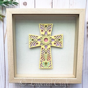 May include: A framed cross with a yellow, pink, and green mandala design. The cross is in a light wood frame with a white mat.
