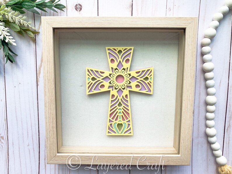 May include: A framed cross with a yellow, pink, and green mandala design. The cross is in a light wood frame with a white mat.