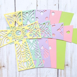 May include: A layered paper craft project featuring a yellow cross with intricate cutouts, surrounded by green, purple, and pink paper squares with various cutouts. The project is on a white wooden background.