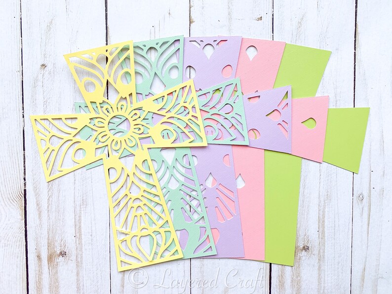 May include: A layered paper craft project featuring a yellow cross with intricate cutouts, surrounded by green, purple, and pink paper squares with various cutouts. The project is on a white wooden background.