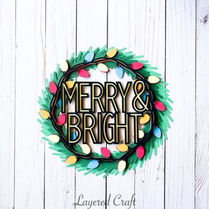 Pu&ograve; includere: Una ghirlanda natalizia a strati con le parole "MERRY & BRIGHT" in oro e nero. La ghirlanda &egrave; decorata con luci colorate e fogliame verde, su uno sfondo di legno bianco.