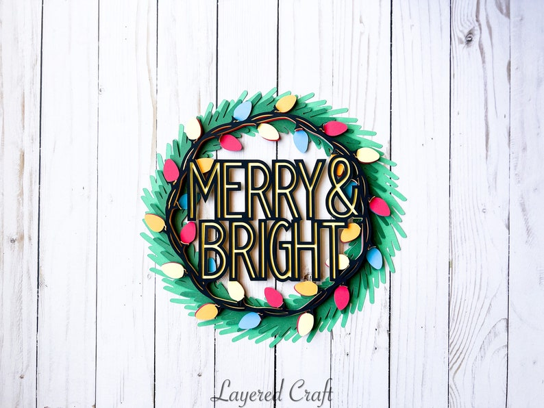 Pu&ograve; includere: Una ghirlanda natalizia a strati con le parole "MERRY & BRIGHT" in oro e nero. La ghirlanda &egrave; decorata con luci colorate e fogliame verde, su uno sfondo di legno bianco.