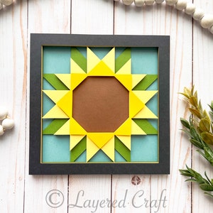May include: A framed paper craft wall art piece depicting a sunflower with yellow, green, and brown paper. The background is a light blue color. The frame is black with a gold inner border. The text "Layered Craft" is visible in the bottom right corner of the image.