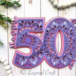 May include: Purple, pink and white layered paper cut-out of the number 50 with a decorative mandala design. The number is on a white wooden background.