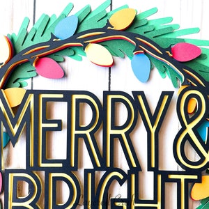Pu&ograve; includere: Una ghirlanda natalizia di carta a strati con le parole "MERRY & BRIGHT". La ghirlanda presenta un fogliame verde, luci colorate e una combinazione di colori nero e giallo. Il design &egrave; festoso e decorativo.