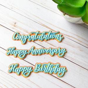 以下が含まれることがあります： 虹色のカラーパターンで重ねられた3つの紙切り。"おめでとう","結婚記念日おめでとう","お誕生日おめでとう"の言葉が筆記体で書かれています。