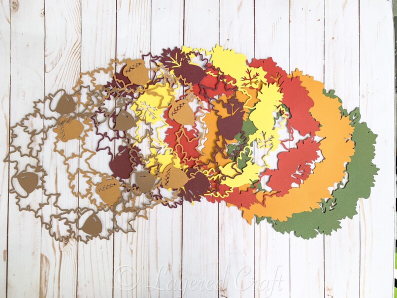 May include: A set of layered paper cutouts in various shades of brown, orange, yellow, and green, featuring leaf and acorn shapes. The cutouts are arranged in a circular pattern, creating a festive autumnal design.