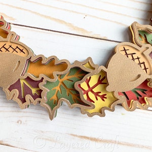 May include: A layered paper craft wreath featuring brown acorns and multi-colored leaves in shades of red, orange, yellow, and green.