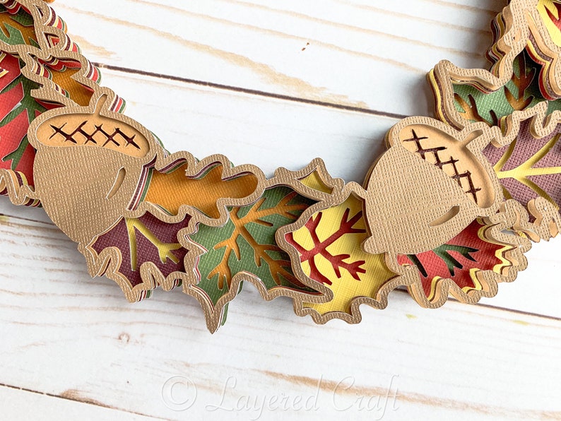 May include: A layered paper craft wreath featuring brown acorns and multi-colored leaves in shades of red, orange, yellow, and green.
