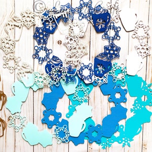 May include: A collection of paper cutouts featuring winter-themed designs. The image includes snowflakes, coffee mugs, and hot chocolate cups in shades of white, blue, and brown. The cutouts are arranged in a circular pattern.