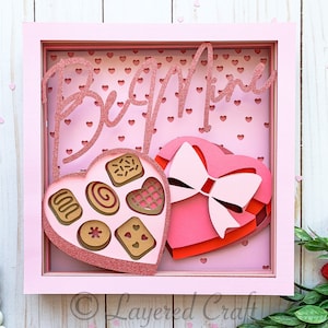 May include: A pink framed shadow box with a pink heart-shaped box with a pink bow and a heart-shaped box of chocolates with the text "Be Mine" on the top.