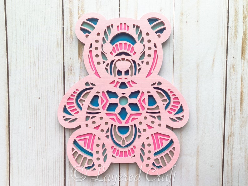 May include: A pink wooden bear with a mandala design in blue, pink, and brown. The bear is cut out in a layered style, creating a 3D effect.