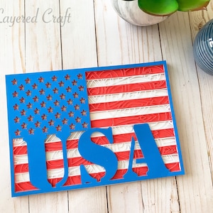 Puede incluir: Una artesanía de papel azul y rojo de la bandera estadounidense con la palabra "USA" recortada en el centro. La bandera está hecha de intrincados diseños de corte de papel.