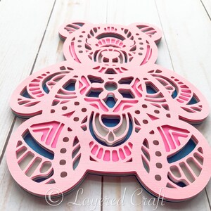 May include: A layered papercraft design of a pink bear with intricate patterns in shades of pink, blue, and beige. The bear is cut out and layered to create a 3D effect.