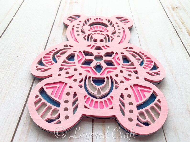 May include: A layered papercraft design of a pink bear with intricate patterns in shades of pink, blue, and beige. The bear is cut out and layered to create a 3D effect.