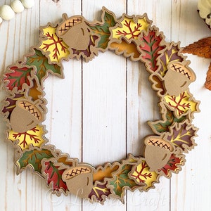 May include: A fall-themed wreath made of layered paper leaves in shades of green, red, yellow, and brown. The wreath is decorated with acorns with stitched details.