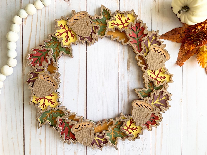 May include: A fall-themed wreath made of layered paper leaves in shades of green, red, yellow, and brown. The wreath is decorated with acorns with stitched details.