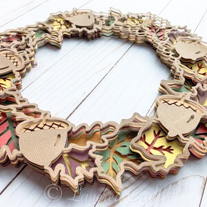 May include: A layered paper craft wreath featuring fall leaves in shades of red, orange, yellow, green, and brown. The wreath is decorated with acorns.