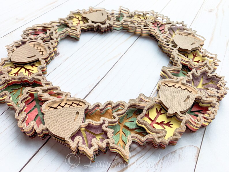 May include: A layered paper craft wreath featuring fall leaves in shades of red, orange, yellow, green, and brown. The wreath is decorated with acorns.