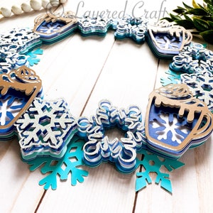 May include: A layered paper wreath featuring a winter theme. The wreath includes blue and silver snowflakes, mugs of hot cocoa, and the text "Layered Craft". The color palette includes shades of blue, silver, and brown.