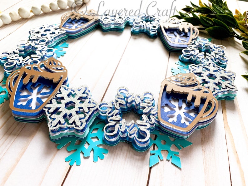 May include: A layered paper wreath featuring a winter theme. The wreath includes blue and silver snowflakes, mugs of hot cocoa, and the text "Layered Craft". The color palette includes shades of blue, silver, and brown.