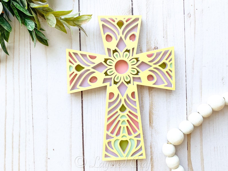 May include: A yellow wooden cross with a pink, green, and purple mandala design. The cross is on a white wooden background.