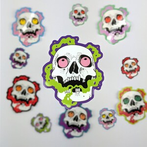 May include: A collection of colourful skull stickers with green, purple, pink and red accents. The stickers feature various skull designs with different expressions and details.