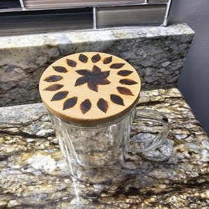 Puede incluir: Una taza de vidrio transparente con una tapa de madera que presenta un diseño de flor de madera quemada. La tapa es redonda y tiene un color marrón oscuro.