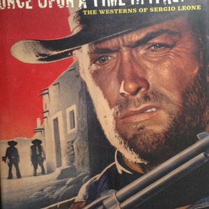 Puede incluir: Una portada de libro con fondo rojo y el título "Érase una vez en el oeste: Los westerns de Sergio Leone". La portada presenta un primer plano de la cara de Clint Eastwood, con un sombrero de vaquero y una pistola en primer plano.