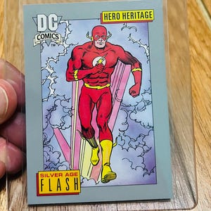 May include: A DC Comics Silver Age Flash trading card in a protective sleeve. The card features a running Flash in a red and yellow costume against a gray background with lightning and the words "Hero Heritage" and "Flash".