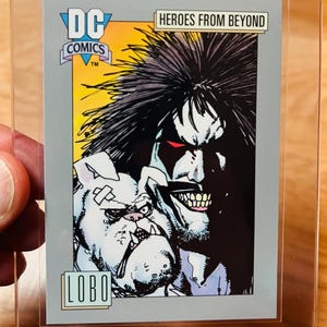 May include: A DC Comics trading card featuring Lobo and a bulldog. The card is in a protective sleeve. The card has the DC Comics logo and the text "HEROES FROM BEYOND" and "LOBO". The artwork is in black, white, and gray.