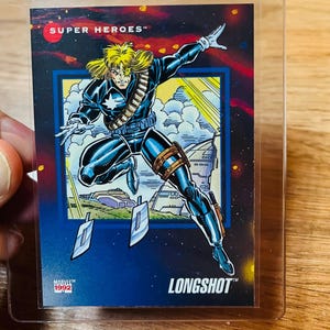 May include: A Longshot Marvel Super Heroes trading card from 1992. The card features a comic book illustration of Longshot in a dynamic pose, with a blue and black costume, blonde hair, and a futuristic background. The card is in a protective sleeve.