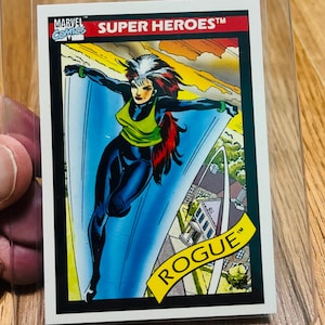 May include: A Marvel Comics Super Heroes trading card featuring Rogue. The card shows Rogue in a dynamic pose with a green and black costume, red hair, and the word "ROGUE" in a yellow banner. The card is in a clear protective sleeve.
