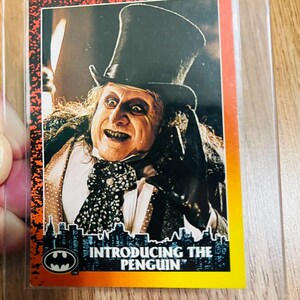 May include: A trading card featuring the Penguin from the Batman franchise. The card has a red border and the text "INTRODUCING THE PENGUIN" at the bottom. The Penguin is depicted in a top hat and tuxedo.