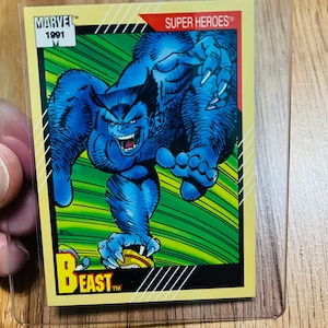 May include: A Marvel Super Heroes trading card from 1991 featuring Beast. The card has a yellow border with the word "BEAST" at the bottom. The Beast character is blue with a fierce expression and is in a protective plastic sleeve.