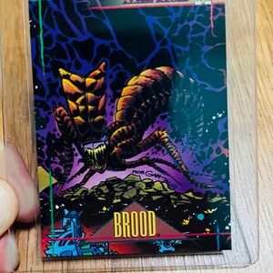 May include: A Marvel Comics trading card featuring the Brood, an alien race. The card has a dark background with a purple and black color scheme. The Brood creature is depicted in shades of yellow, orange, and brown. The word "BROOD" is prominently displayed.