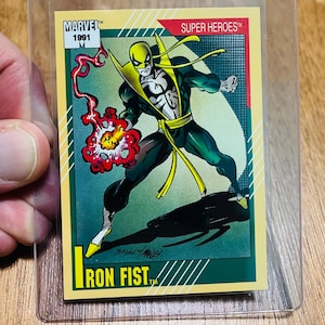 May include: A 1991 Marvel Super Heroes Iron Fist trading card in a protective sleeve. The card features a dynamic illustration of Iron Fist in a fighting pose, with a red energy blast. The card has a yellow border and the text "IRON FIST".