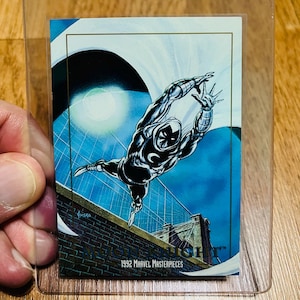 May include: A 1992 Marvel Masterpieces trading card featuring the character Moon Knight in a dynamic pose. The card is encased in a clear plastic holder. The background shows a bridge and a blue sky.