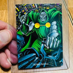 May include: A trading card featuring Doctor Doom from the 1992 Marvel Masterpieces series. The card shows Doctor Doom in a green cloak and silver armor, with a cityscape background. The card is encased in a clear plastic card saver.