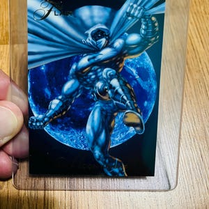 May include: A trading card featuring a muscular character in a white and blue costume, set against a blue moon backdrop. The card is encased in a clear plastic sleeve. The word "Flair" is visible on the card.