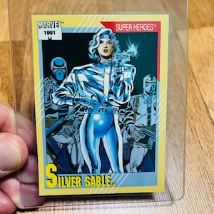 May include: A white ceramic mug with a black handle and the text "But First, Coffee" in a bold, black font. The mug is set against a blurred background, suggesting a cozy setting.