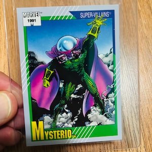 May include: A 1991 Marvel Super-Villains trading card featuring Mysterio. The card shows a green and purple costumed character with a fishbowl helmet, set against a blue sky and purple cape. The card is in a clear protective sleeve.