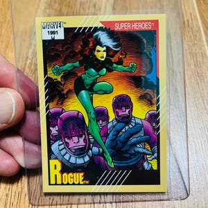 May include: A 1991 Marvel Super Heroes trading card featuring Rogue in a green and black costume, with white hair and a yellow background. The card also shows a group of purple robots and the word "Rogue".
