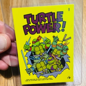 May include: A yellow trading card featuring the Teenage Mutant Ninja Turtles with the words "TURTLE POWER!" in purple. The card shows the turtles in action poses with their weapons. The card is in a protective sleeve.