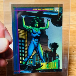 May include: A She-Hulk trading card featuring a green-skinned superheroine in a blue bodysuit lifting a vehicle. The card has the text "SUPER HEROES" and "SHE-HULK" in yellow. The background shows a cityscape and a dark green and black structure.